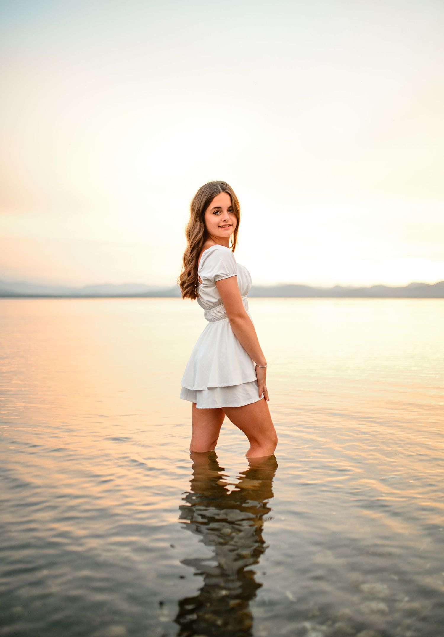 Person in white dress stands in shallow water during golden hour at a serene beach setting.