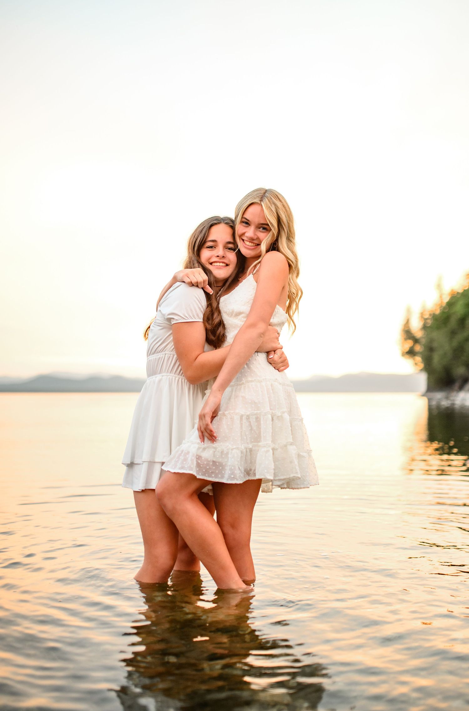 Two friends in white dresses embrace while standing in lake water at golden hour.