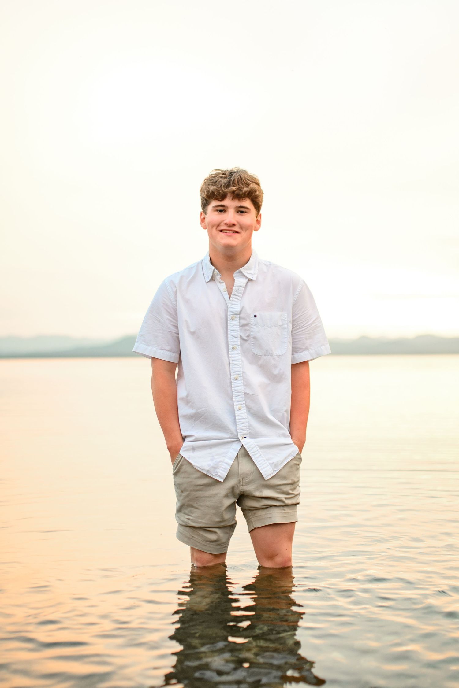 Young person standing in shallow water wearing a light button-down shirt and khaki shorts at sunset.