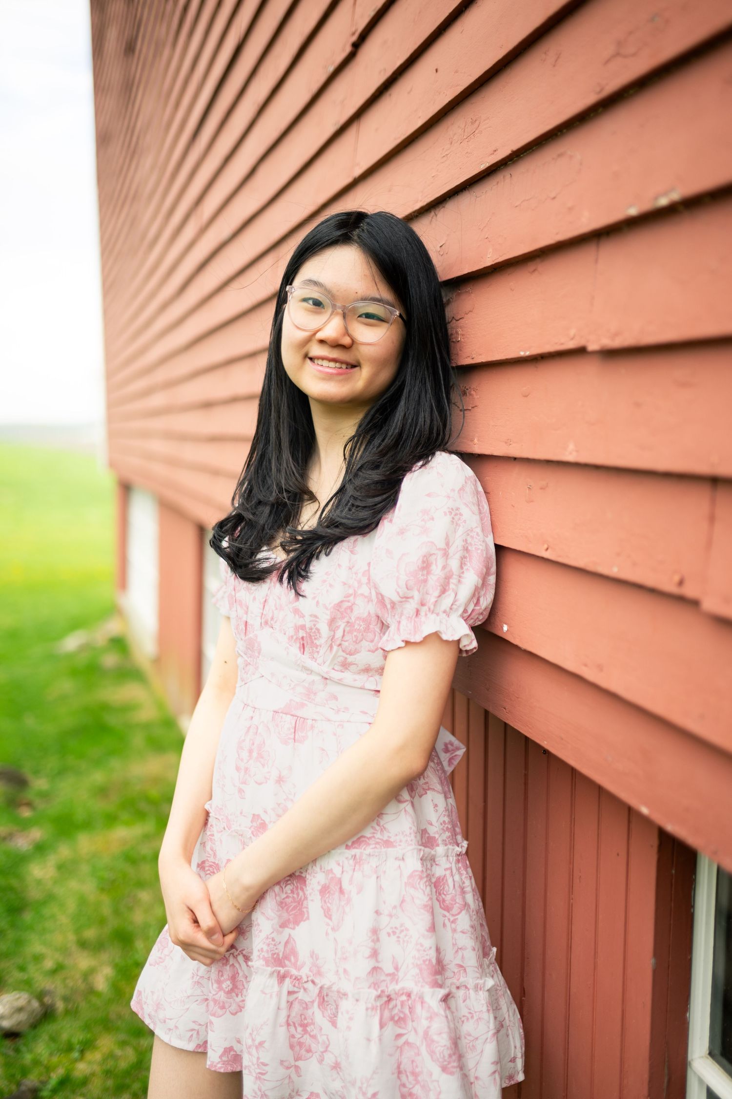Person in a pink floral dress poses against a rustic red barn wall with green grass in the background.