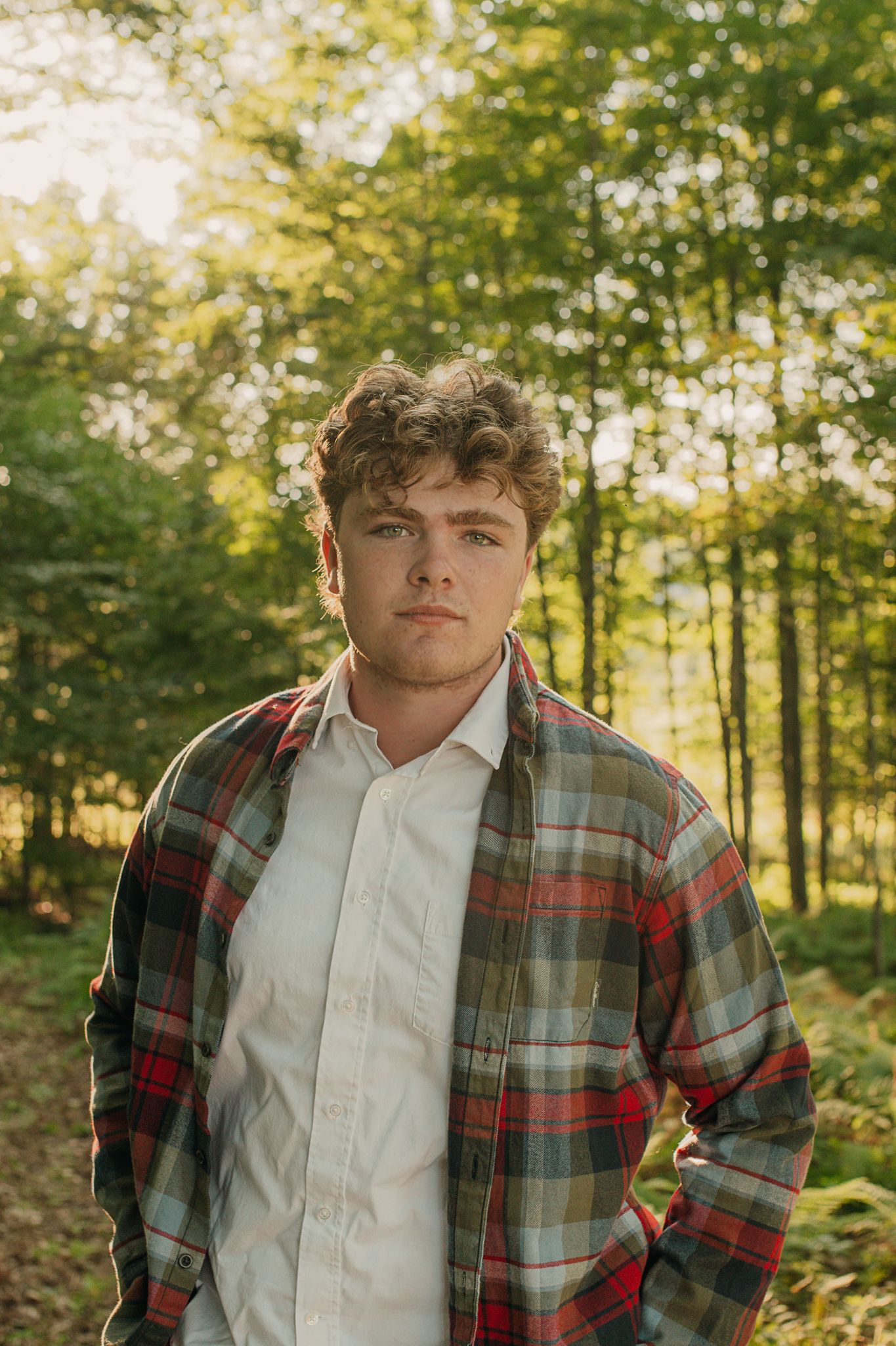 A casual outdoor photoshoot featuring someone in a plaid flannel shirt and white button-down against a sunlit forest backdrop.