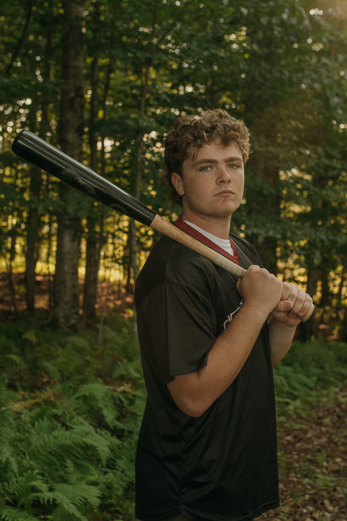 Someone in a black t-shirt poses with a shotgun in a wooded setting during golden hour.
