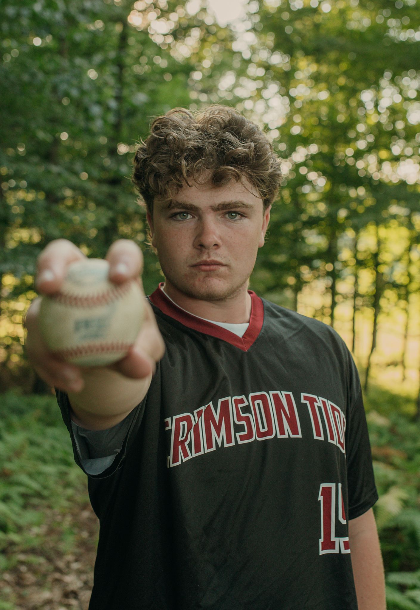 A baseball player in a crimson team uniform holds out a baseball in a wooded outdoor setting.
