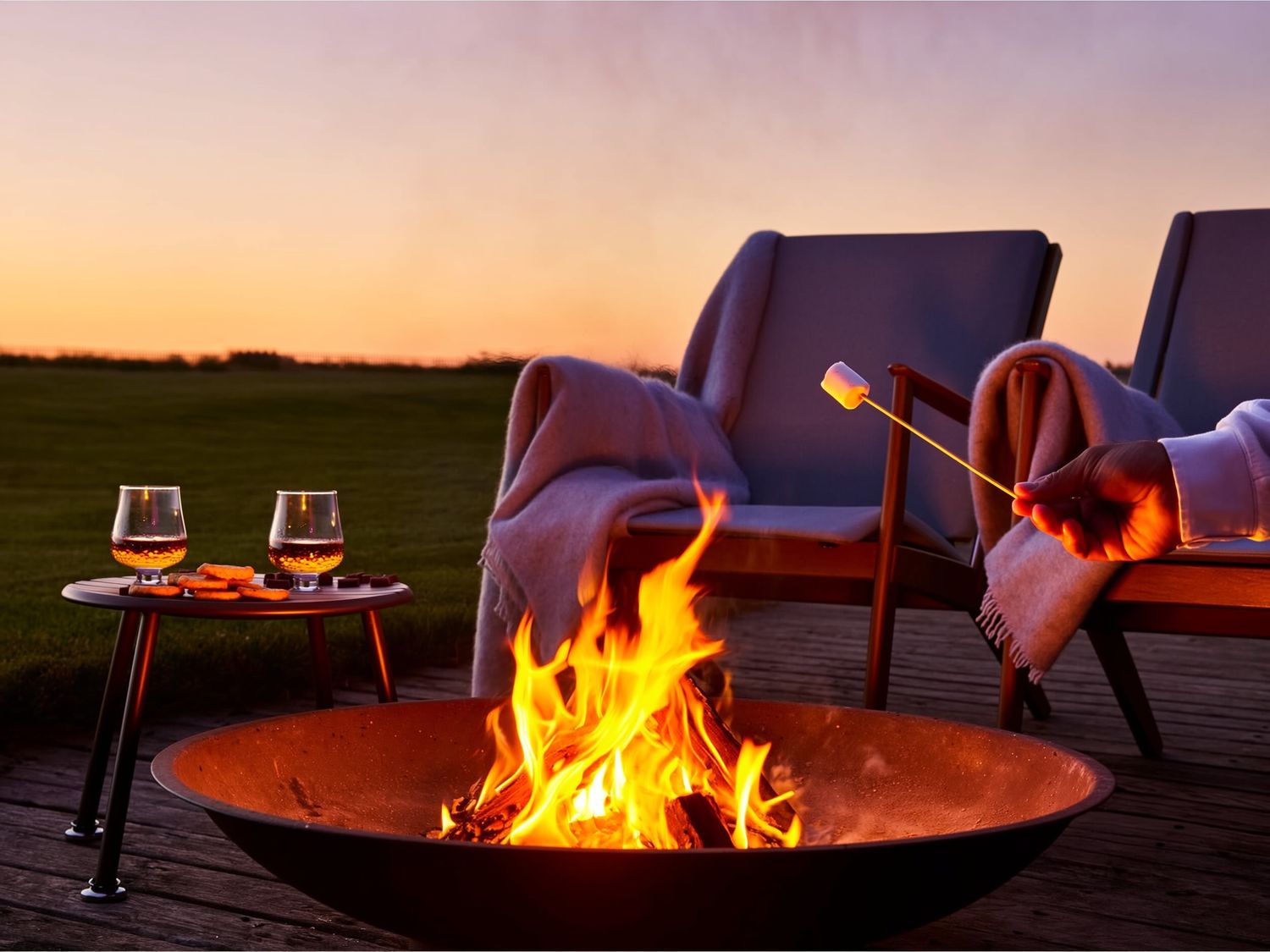 Fire pit glowing at sunset with wine glasses and seating visible in the background.