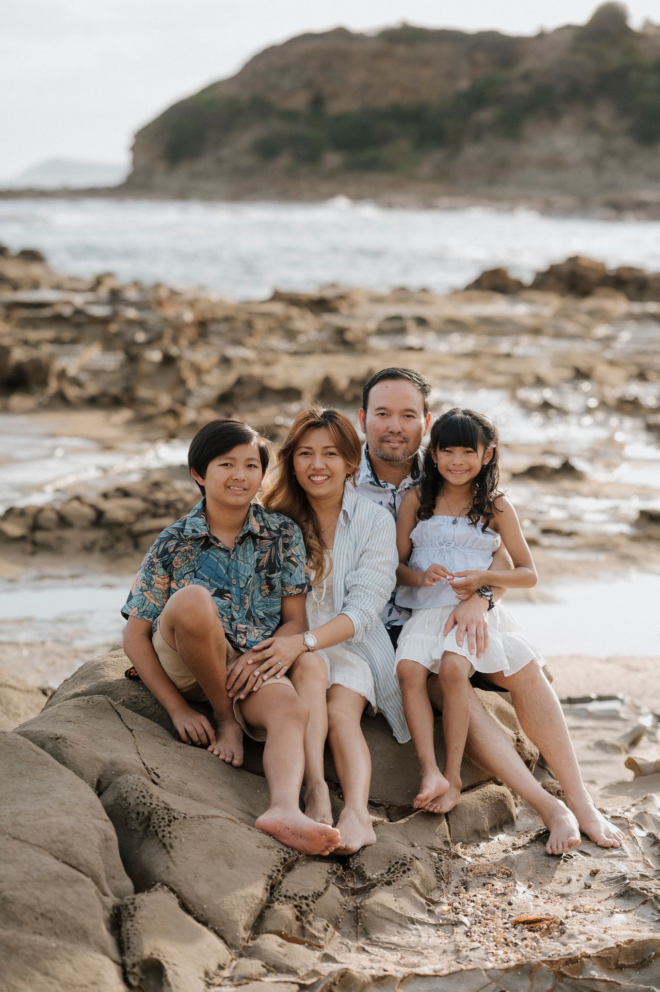 A family sits together barefoot on coastal rocks during sunset at a rocky beach with ocean and cliffs in the background.