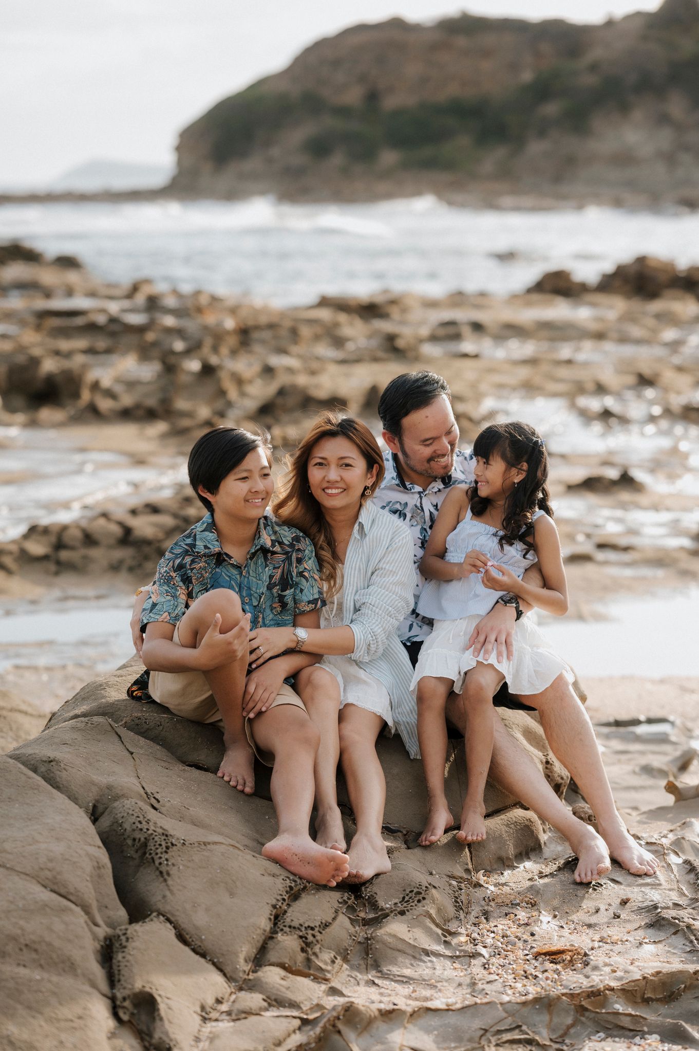 A family sits together on coastal rocks at the beach during golden hour, surrounded by rugged shoreline and ocean waves.