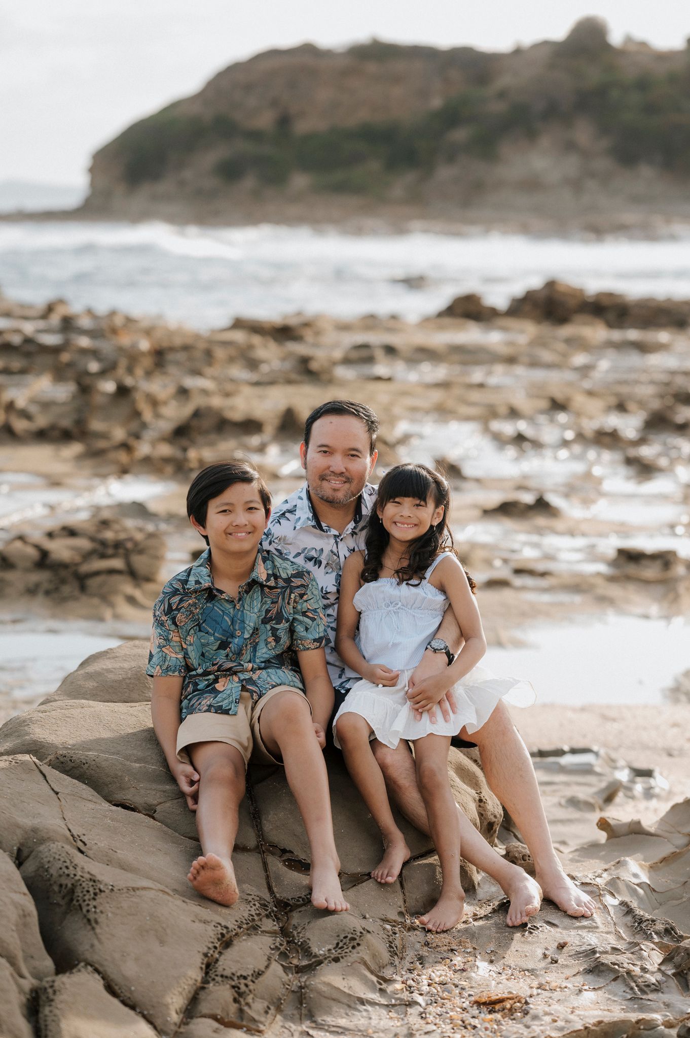 A family sits together on rocky coastal terrain with waves and cliffs visible in the background during a beach photoshoot.