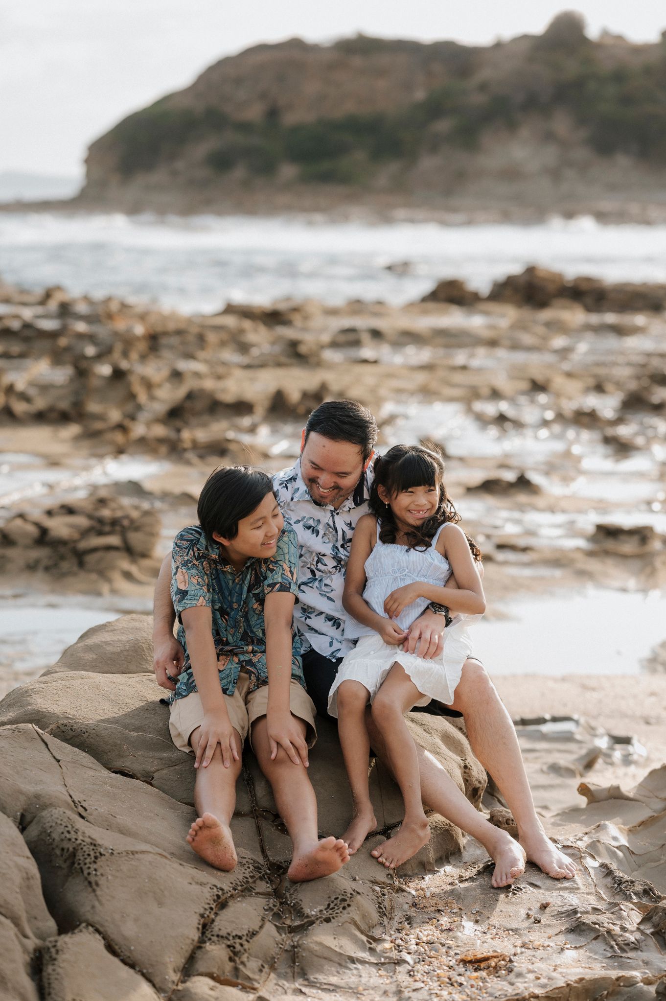 A family sits together on coastal rocks at the beach during golden hour with ocean waves and cliffs in the background.