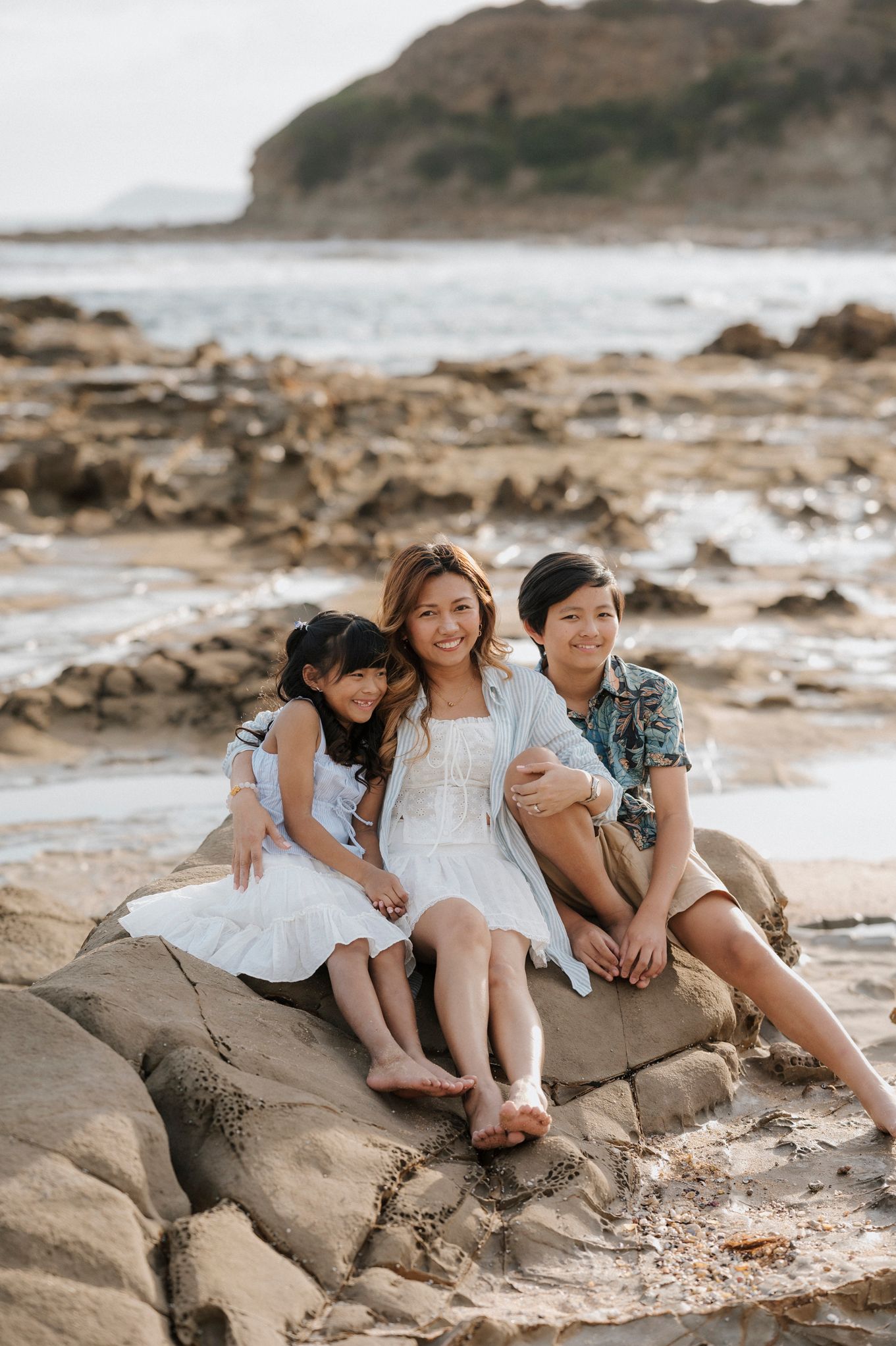 Three people in casual outfits sit together on coastal rocks at the beach with cliffs and ocean in the background.
