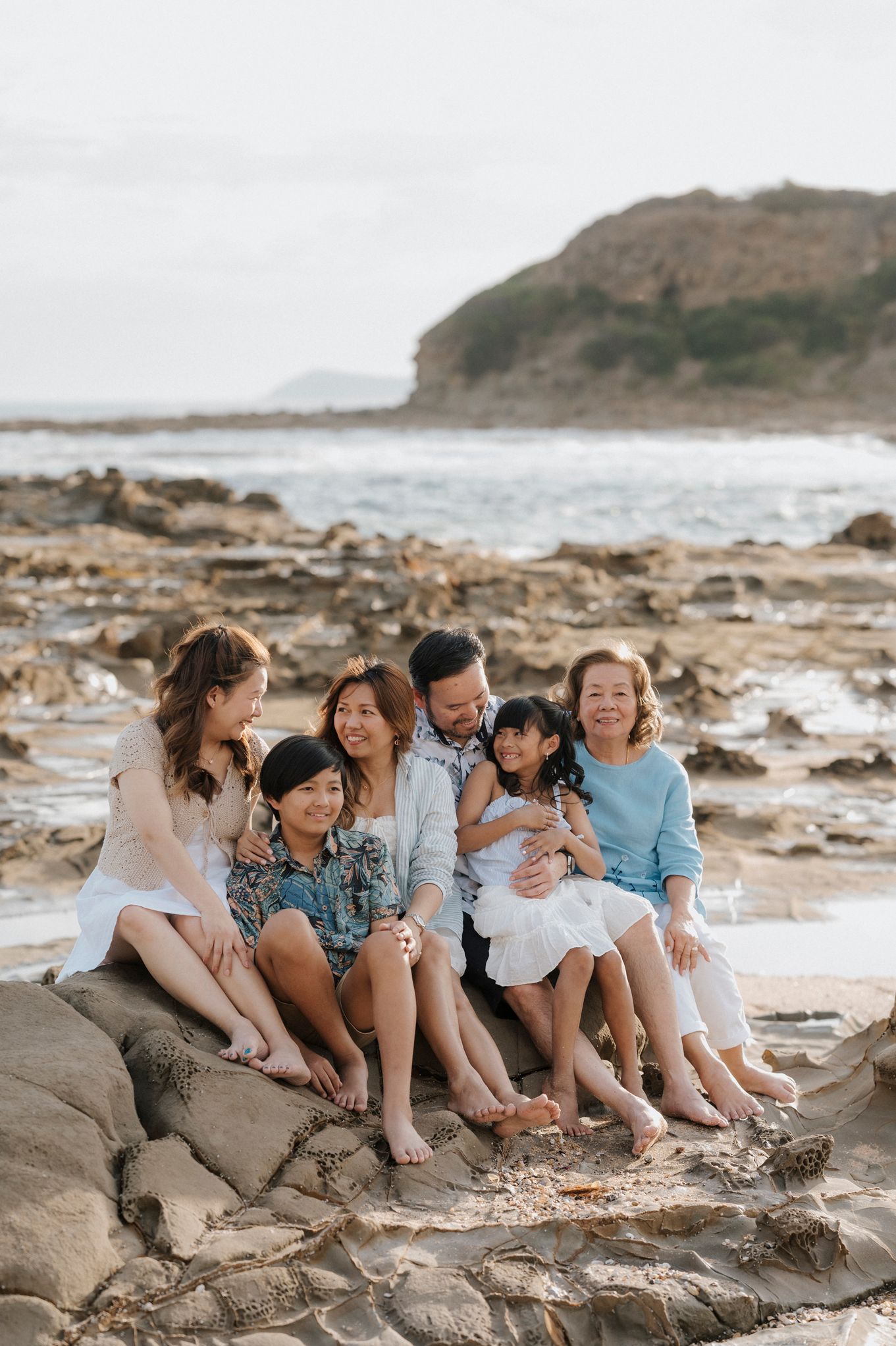 Family gathering on rocky coastal beach with cliffside and ocean view on overcast day.