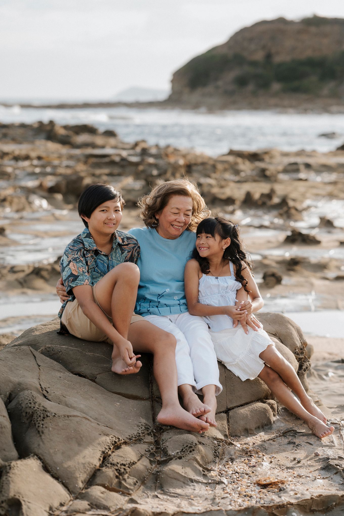 Three people sit together on coastal rocks at sunset, sharing laughs near the ocean's edge.