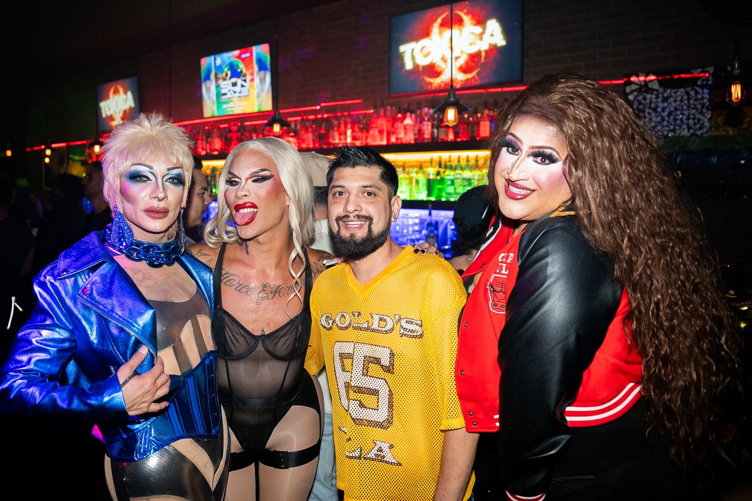 Drag queen in a short blonde wig, one in a long blonde wig stand next to a man in a yellow jersey and a drag queen with dark curly hair.