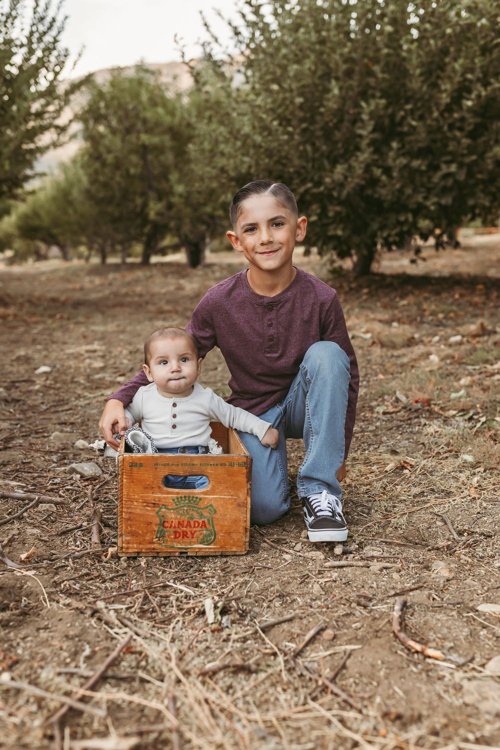A child and baby pose for a portrait with a vintage wooden crate in an outdoor setting.