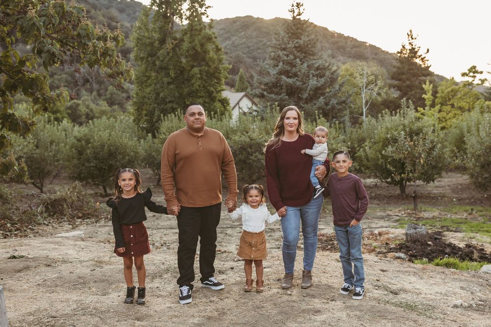 A family of five stands together outdoors on a dirt path with mountains and trees in the background during golden hour.