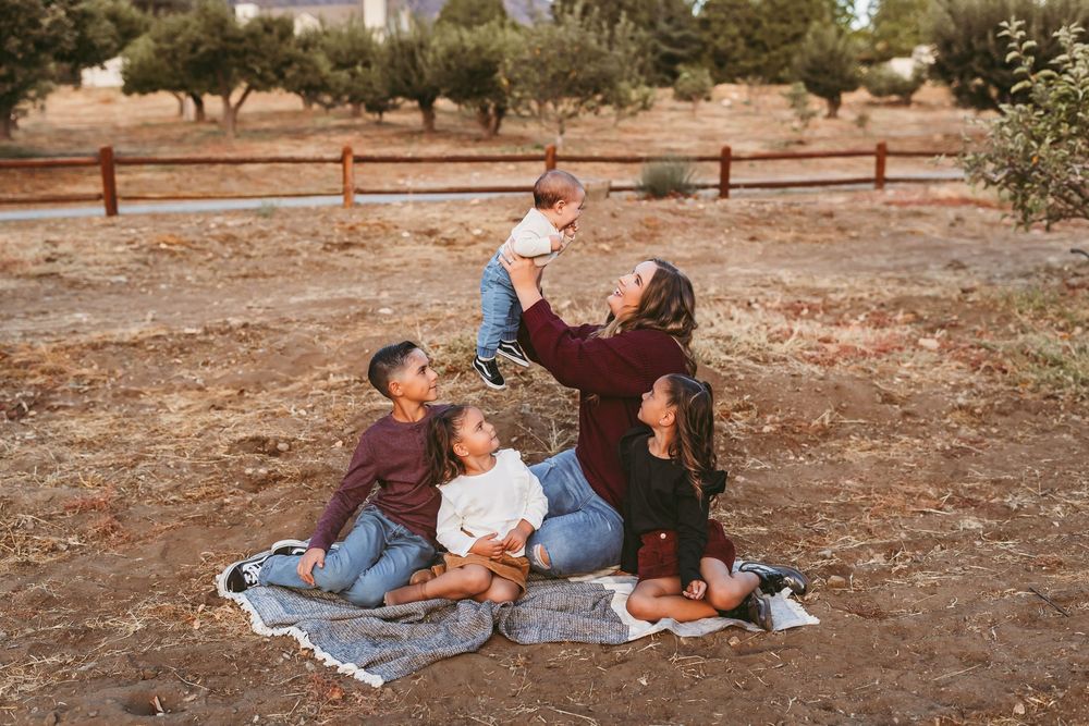Family having a picnic together on a blanket in a rustic outdoor setting with wooden fence and dry ground.