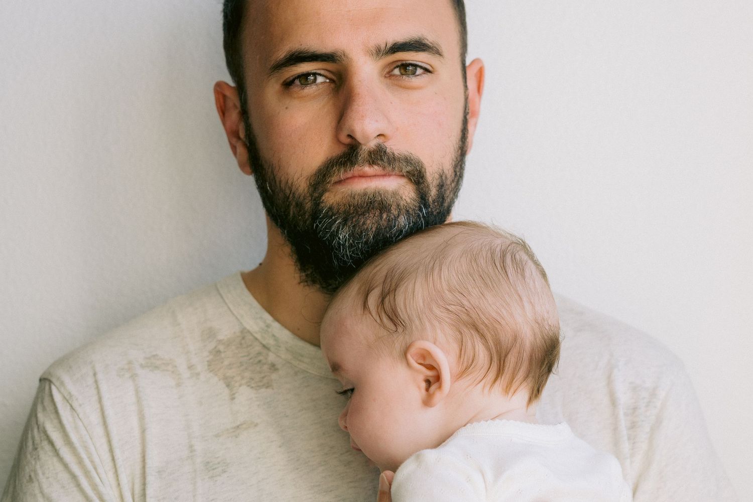 A close-up portrait with father in a beige shirt tenderly holding a baby against his shoulder.