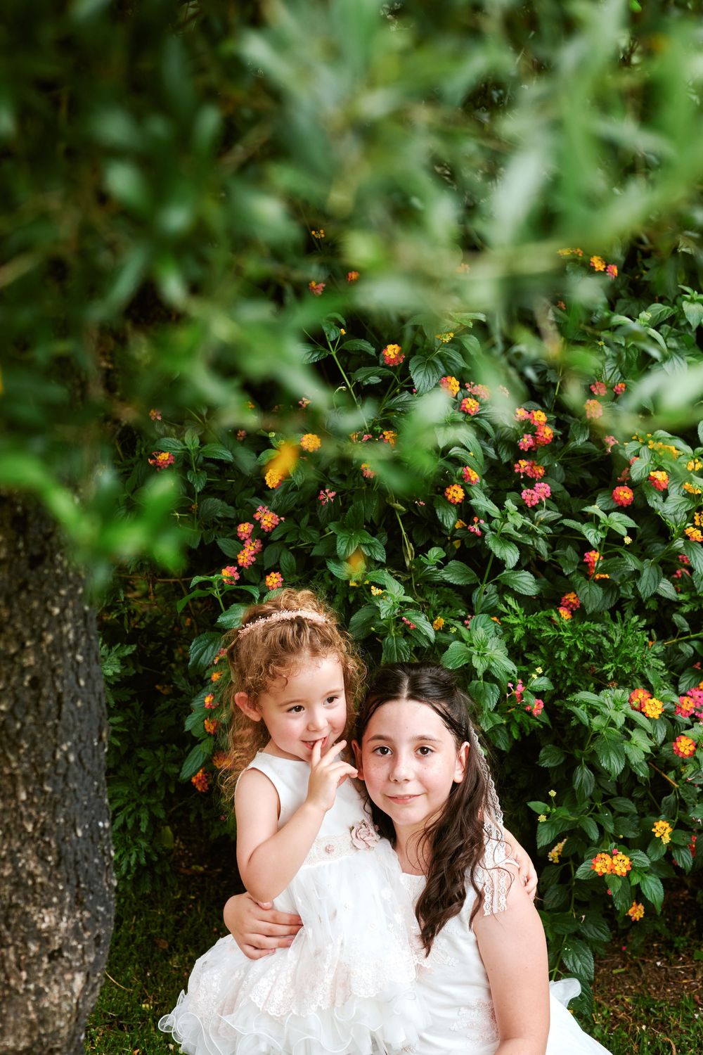 Two young girls in white dresses posing together in front of lush flowering garden.