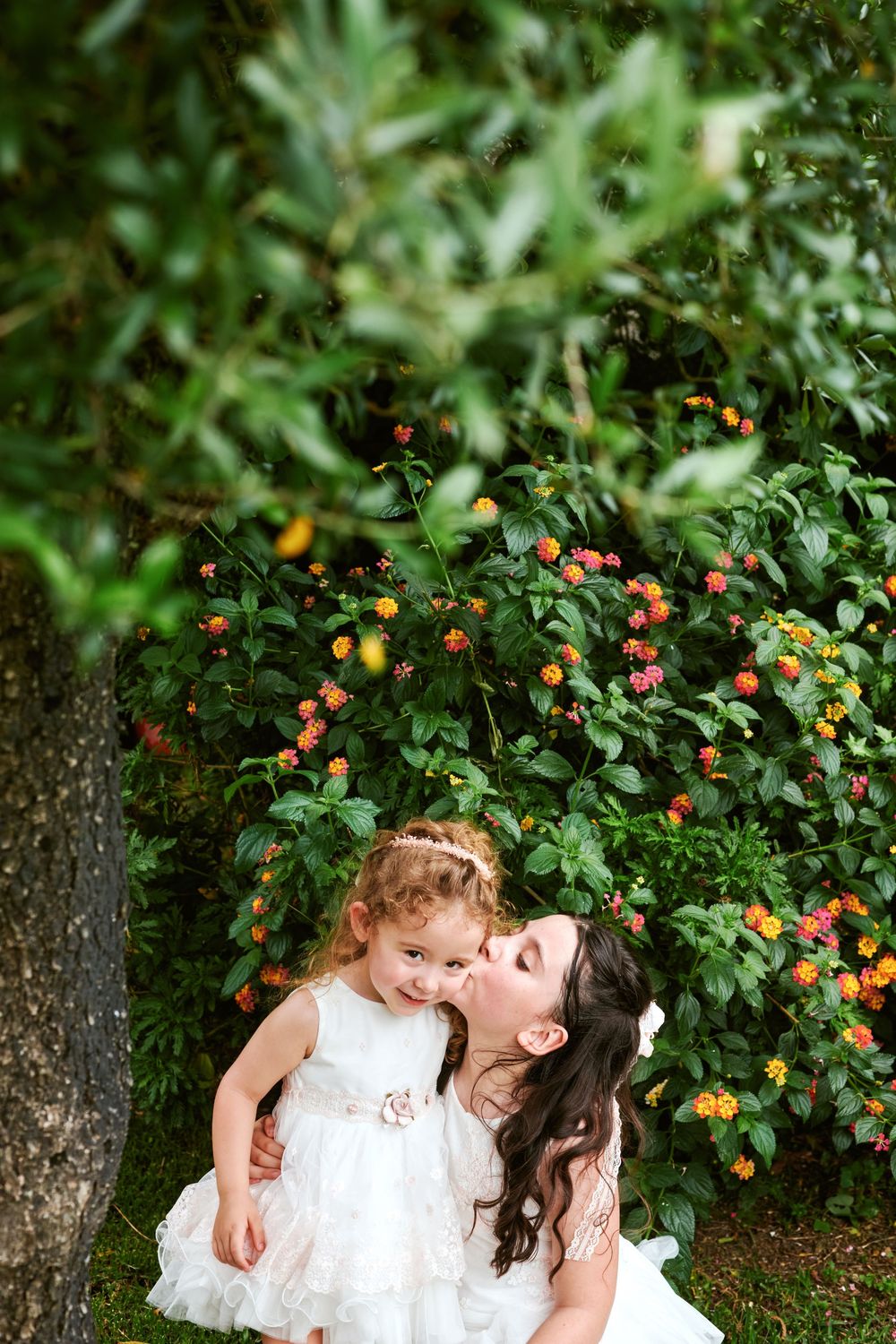 Mother kissing young daughter in white dress while sitting in front of colorful garden.