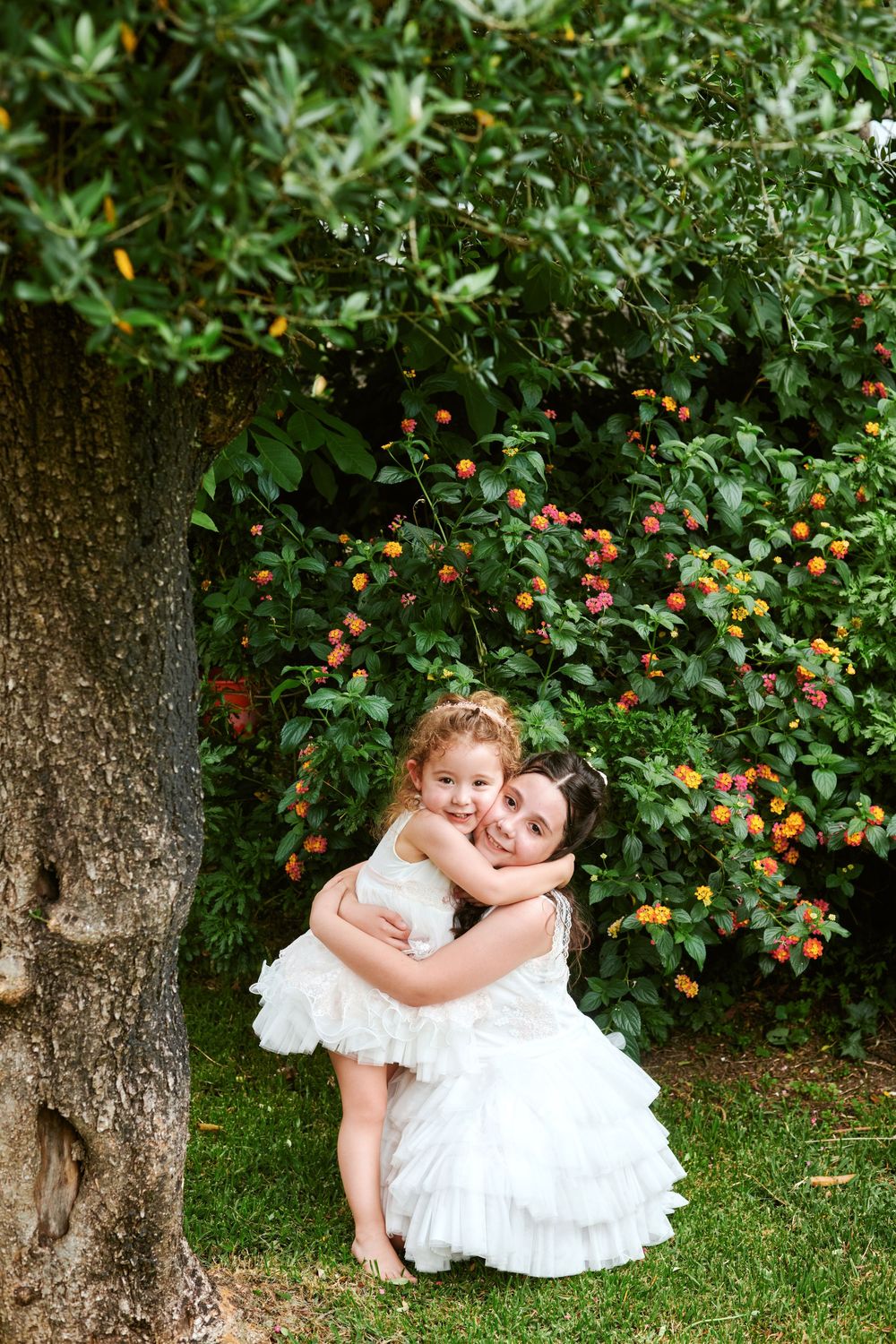 Mother in white dress hugging young girl near flowering shrubs in garden.