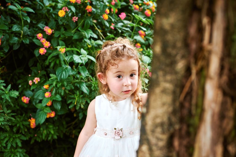 Cute little girl in white dress standing near tree with colorful flowers.