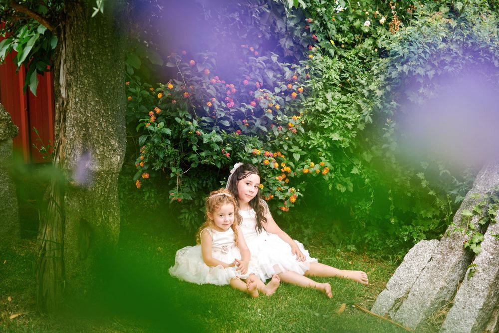 Two girls in white dresses sitting together under tree in lush garden.