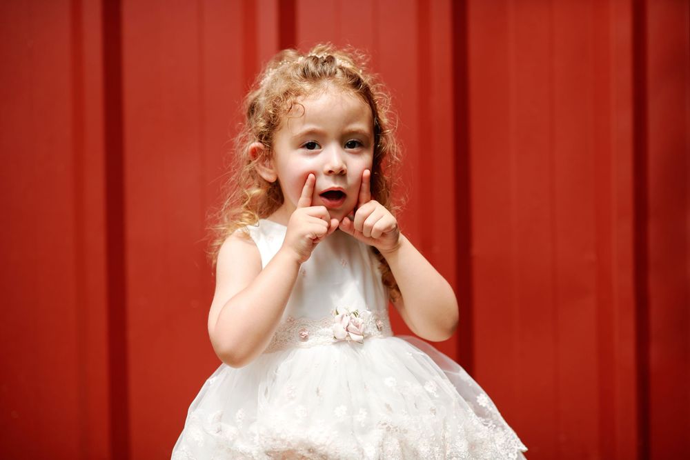 Adorable curly-haired girl in white dress posing against red wall.