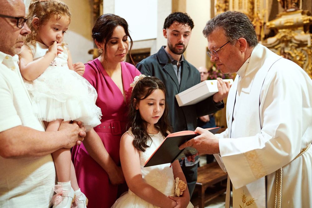 Priest reading from book during baptism ceremony with family including baby in white dress.