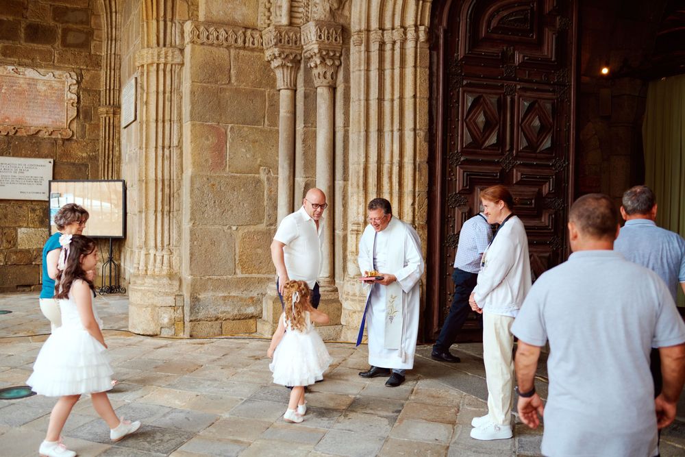 Priest greeting children in white dresses at ornate stone church entrance during religious ceremony.