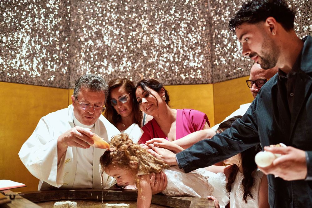 Priest performing baptism water ritual on infant surrounded by smiling family members.