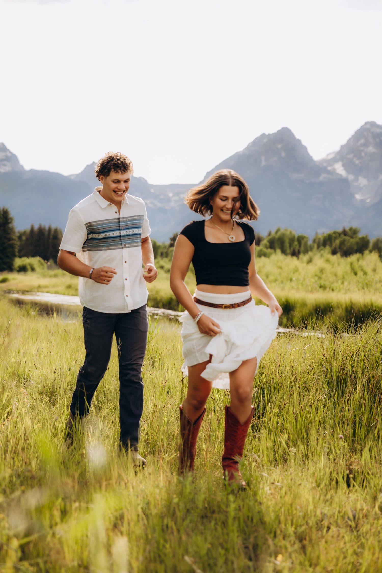 A couple playfully walks through tall grass with mountains visible in the background on a sunny day.
