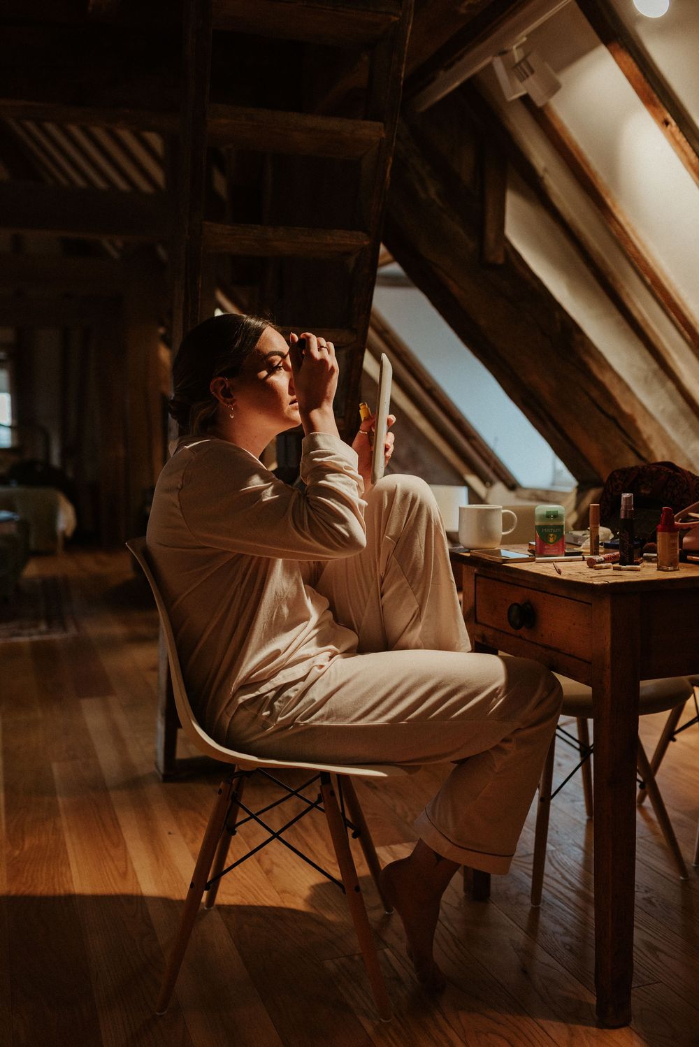 Person in white outfit applying makeup in attic room with warm lighting.