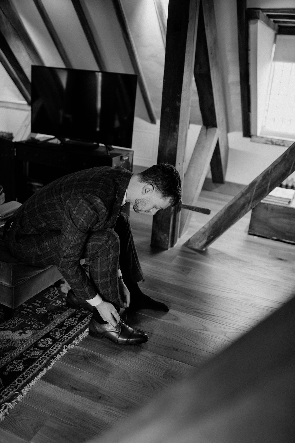 Person in plaid shirt tying shoes on hardwood floor under exposed wooden beams.