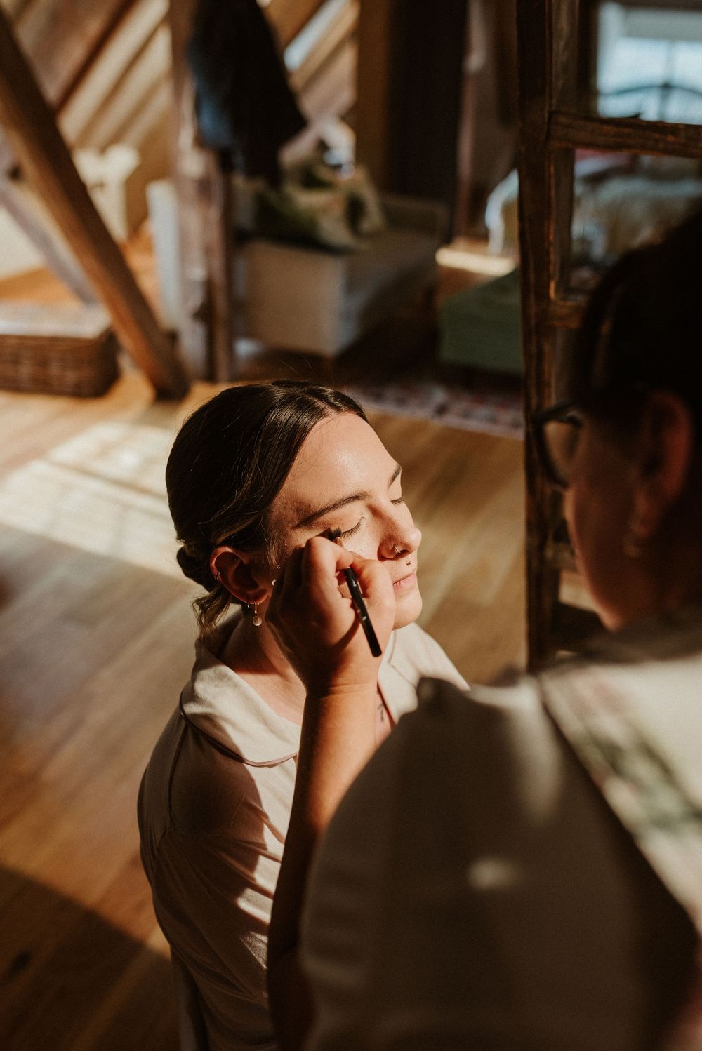 A makeup artist applies eyeshadow to a bride during wedding preparations in warm natural lighting.