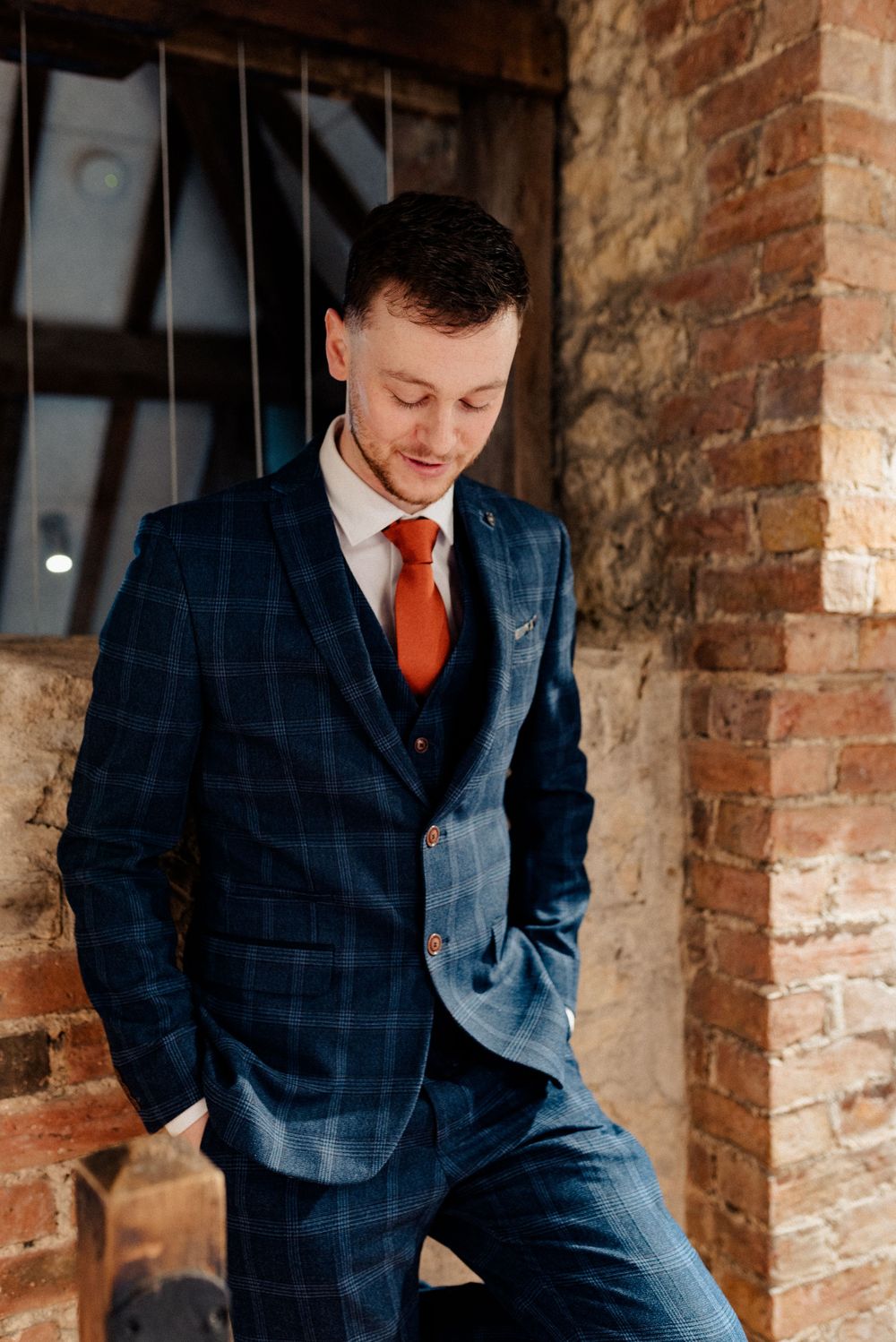 Figure in navy checkered suit with red tie leaning against rustic brick wall.