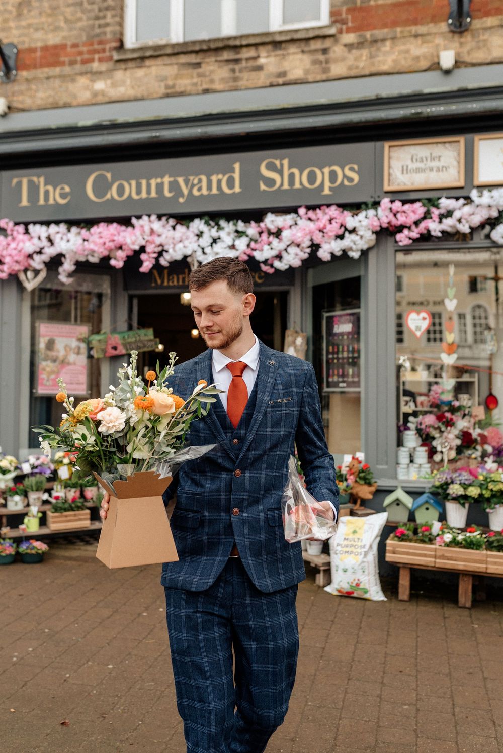 Man in blue checkered suit carrying flowers outside The Courtyard Shops with cherry blossoms.