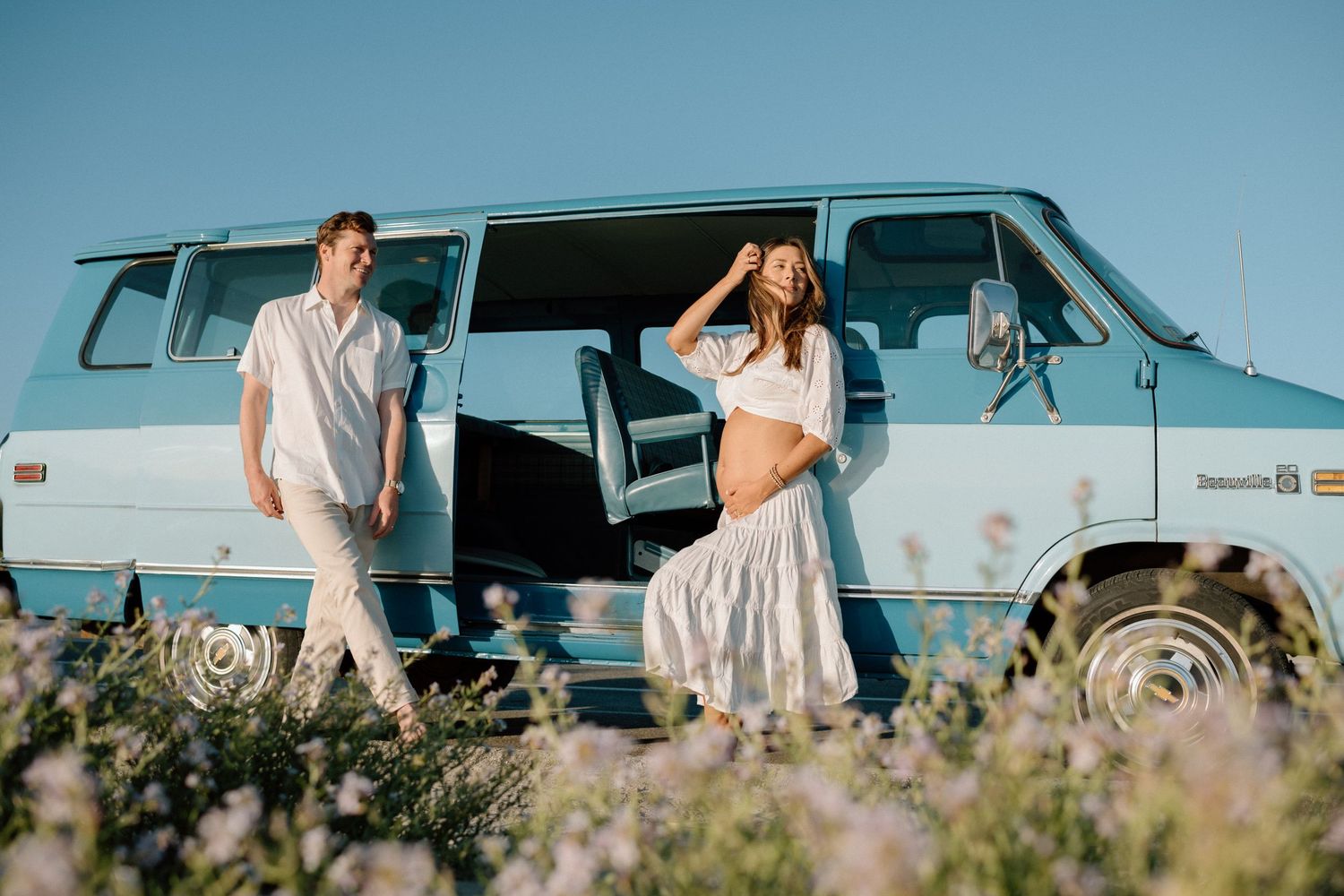 Couple in white outfits standing by a vintage blue van in a wildflower field against blue sky backdrop.