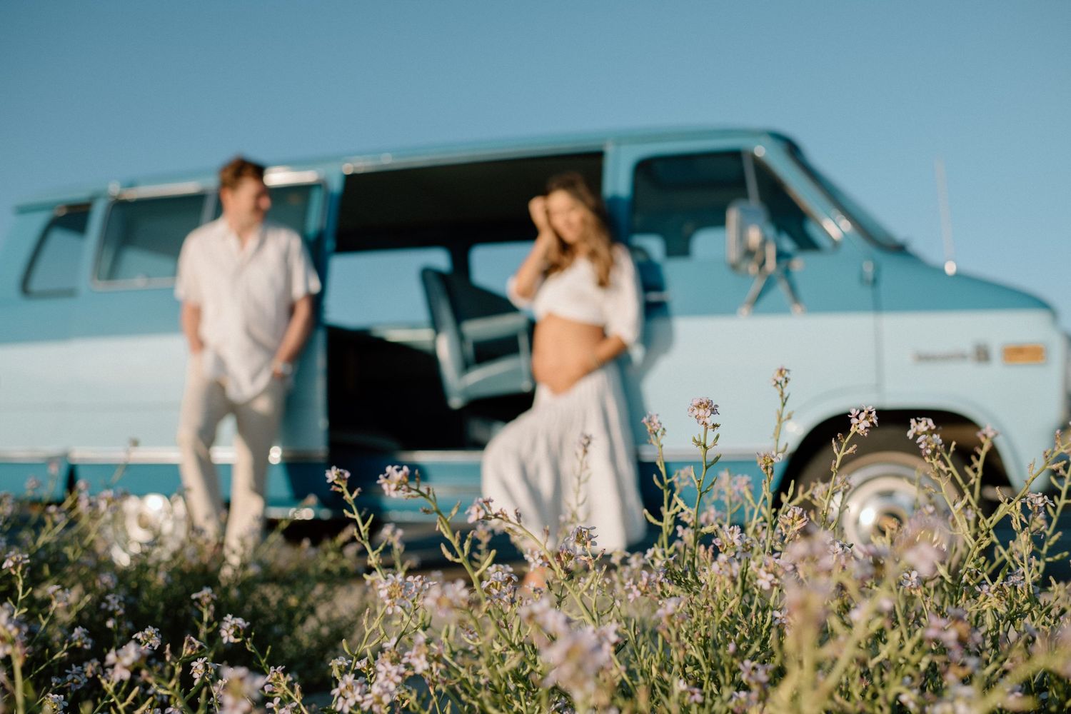 A vintage blue van parked in a wildflower field on a sunny day with two people enjoying the summer scenery.