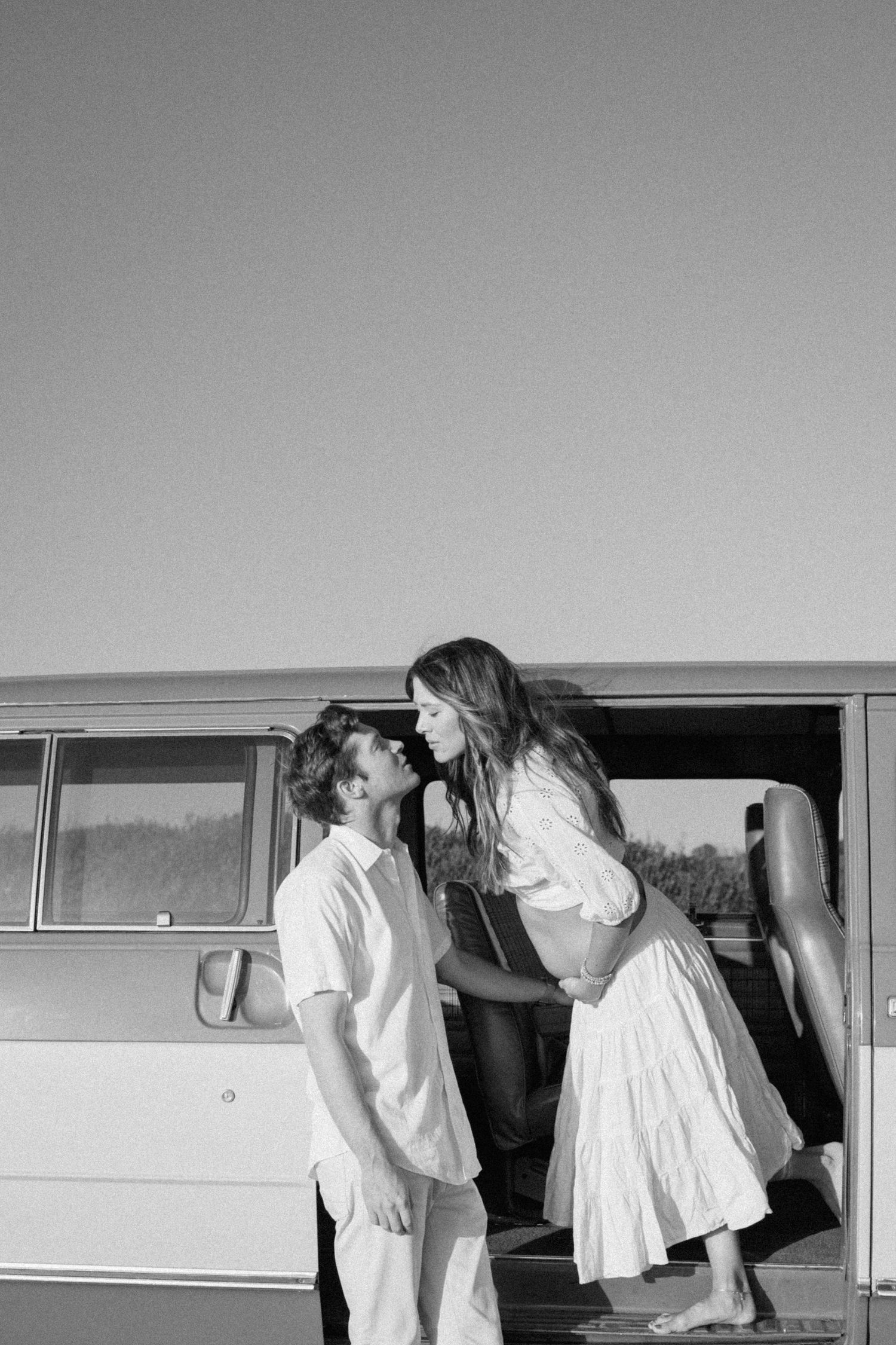 Black and white photo of a romantic vintage scene with a couple by a retro bus, dressed in light summer attire.