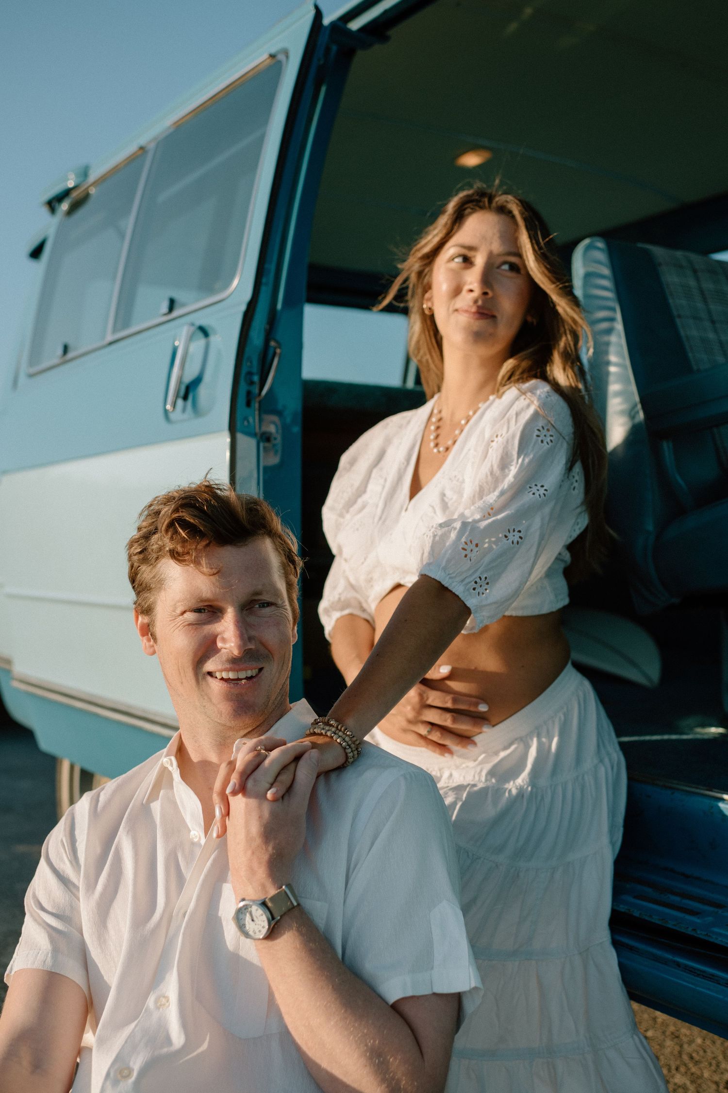 A couple dressed in white poses together near a blue vintage van on a sunny day.