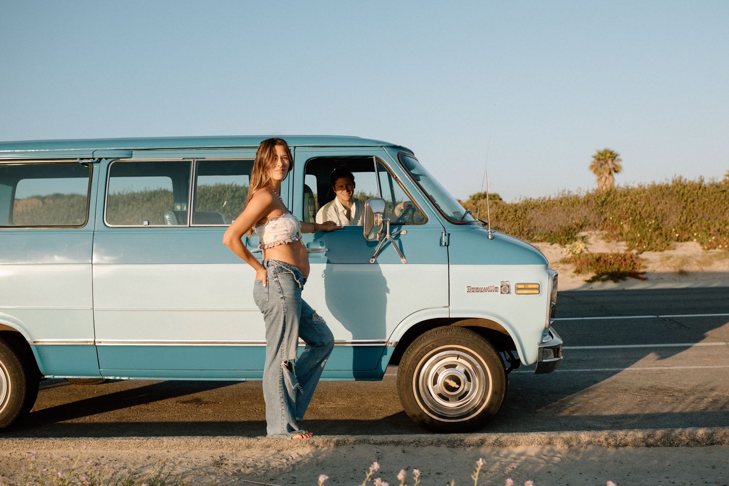 Woman in denim sits beside a vintage blue station wagon on a coastal road at sunset.