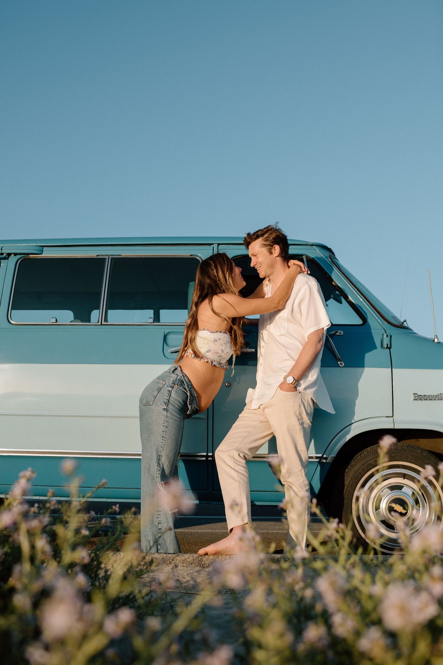 A romantic couple shares a tender moment by a blue vintage van against a clear summer sky.