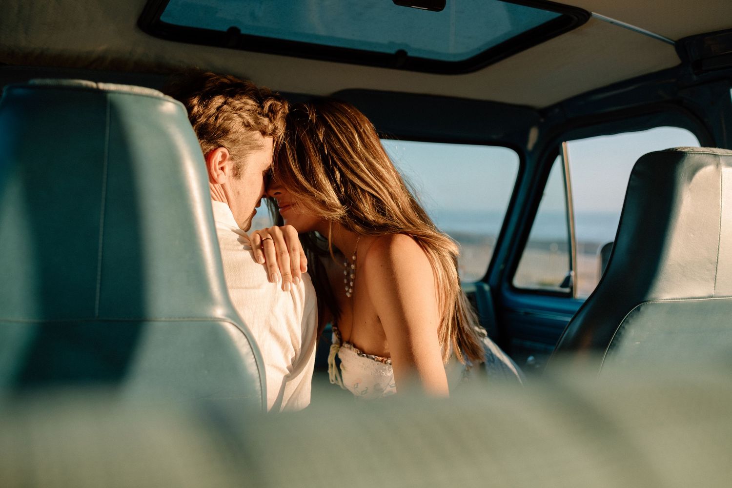 Two people enjoying a romantic moment inside a car at sunset, viewed through the vehicle's windows and glass.