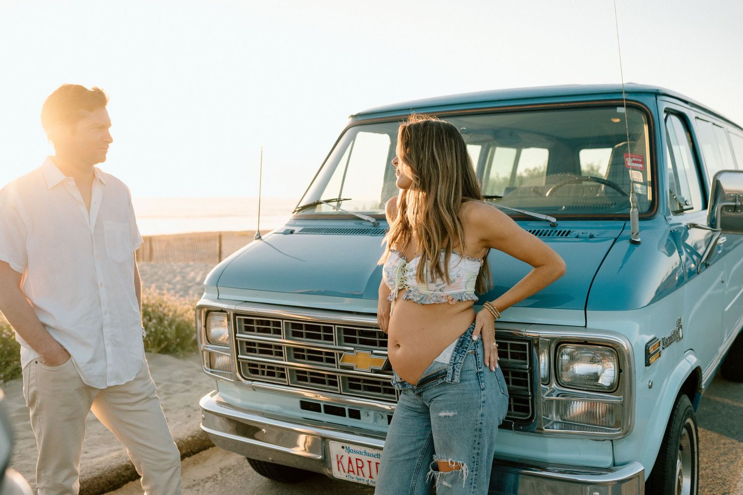 Romantic couple leaning on classic blue vintage car at sunset in beach parking lot with other cars visible.