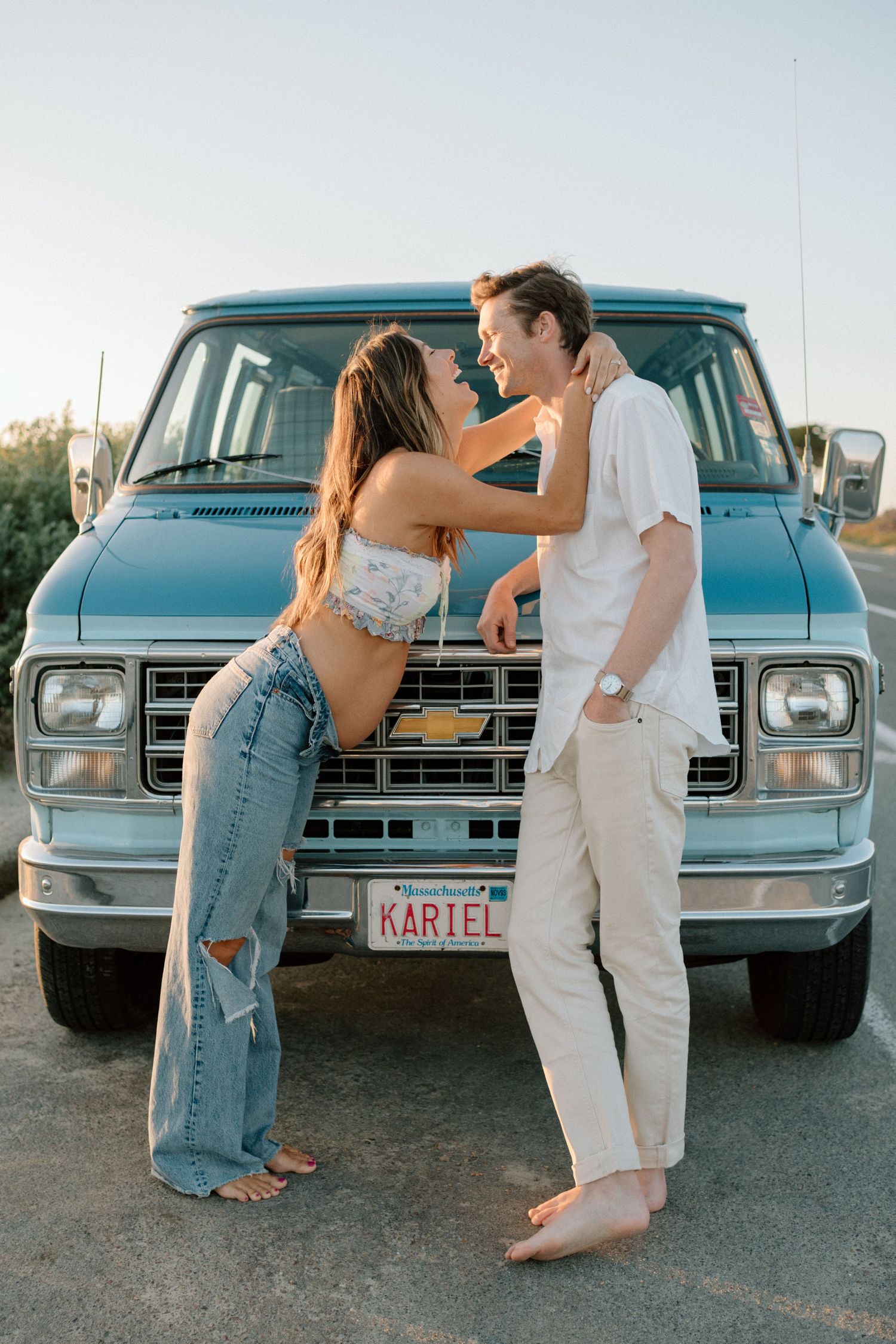 A couple stands intimately in front of a classic blue van wearing casual summer attire.