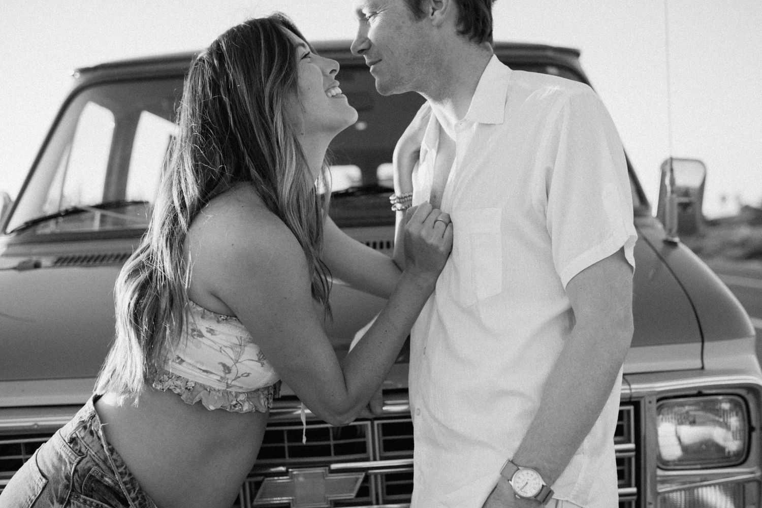 A romantic black and white moment between a couple leaning against a vintage car.