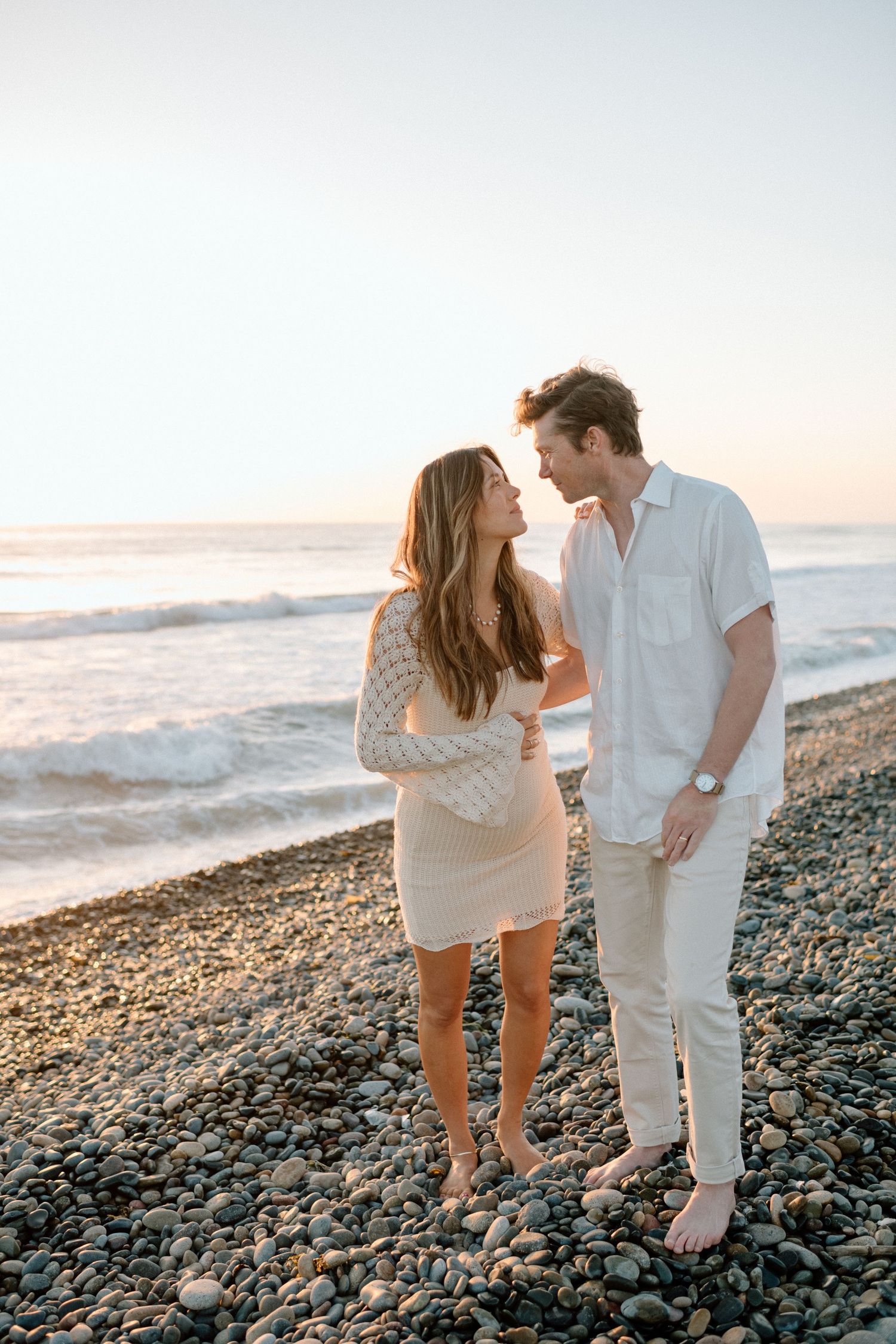 A couple share a romantic moment on a rocky beach at sunset.