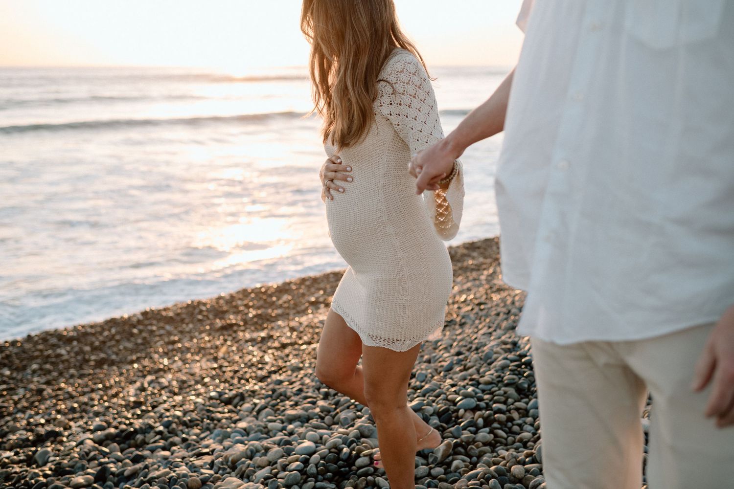 A couple walks hand in hand on a rocky beach at sunset.