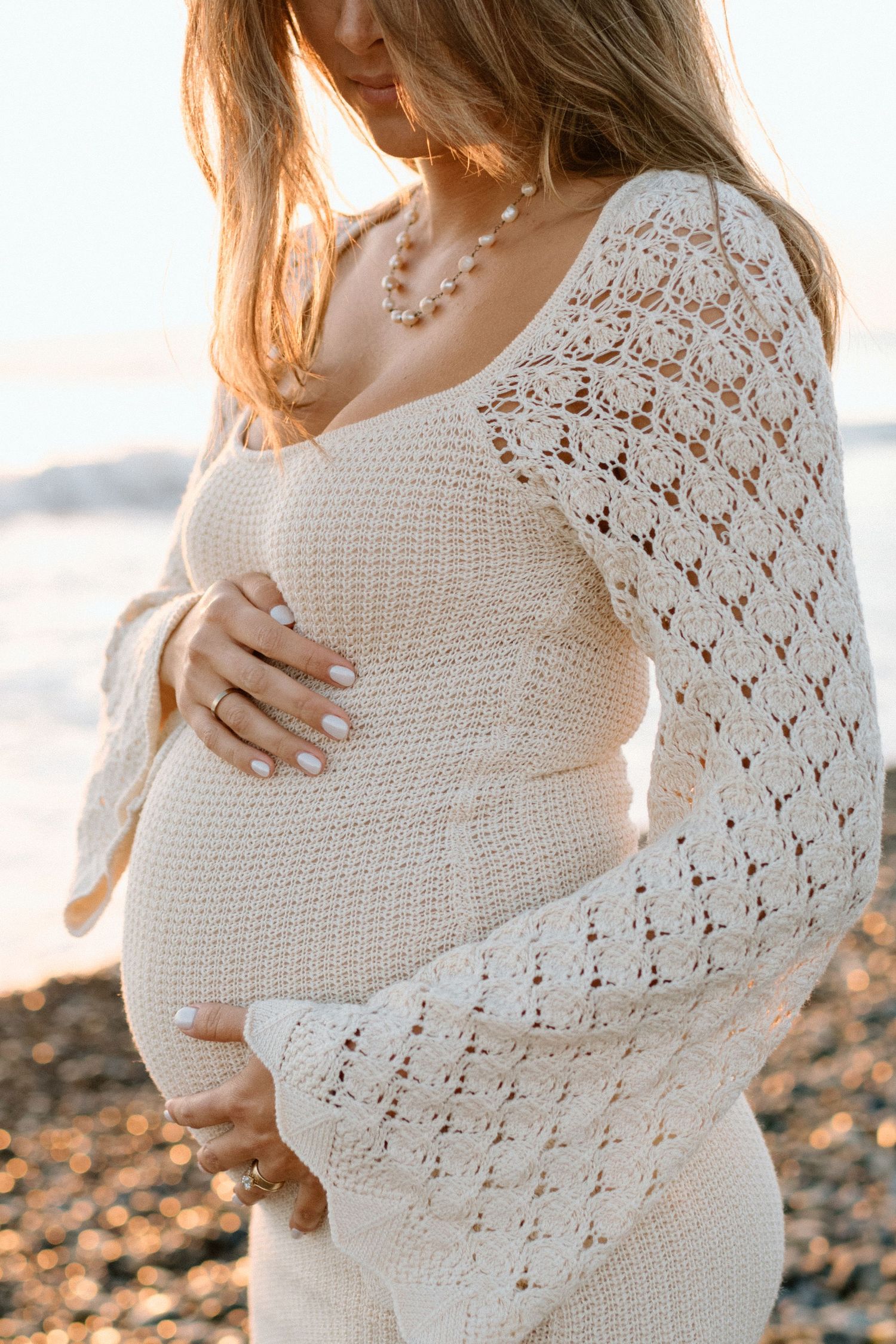 A close-up shot of an expecting mother in a white crochet dress cradling her baby bump at sunset.