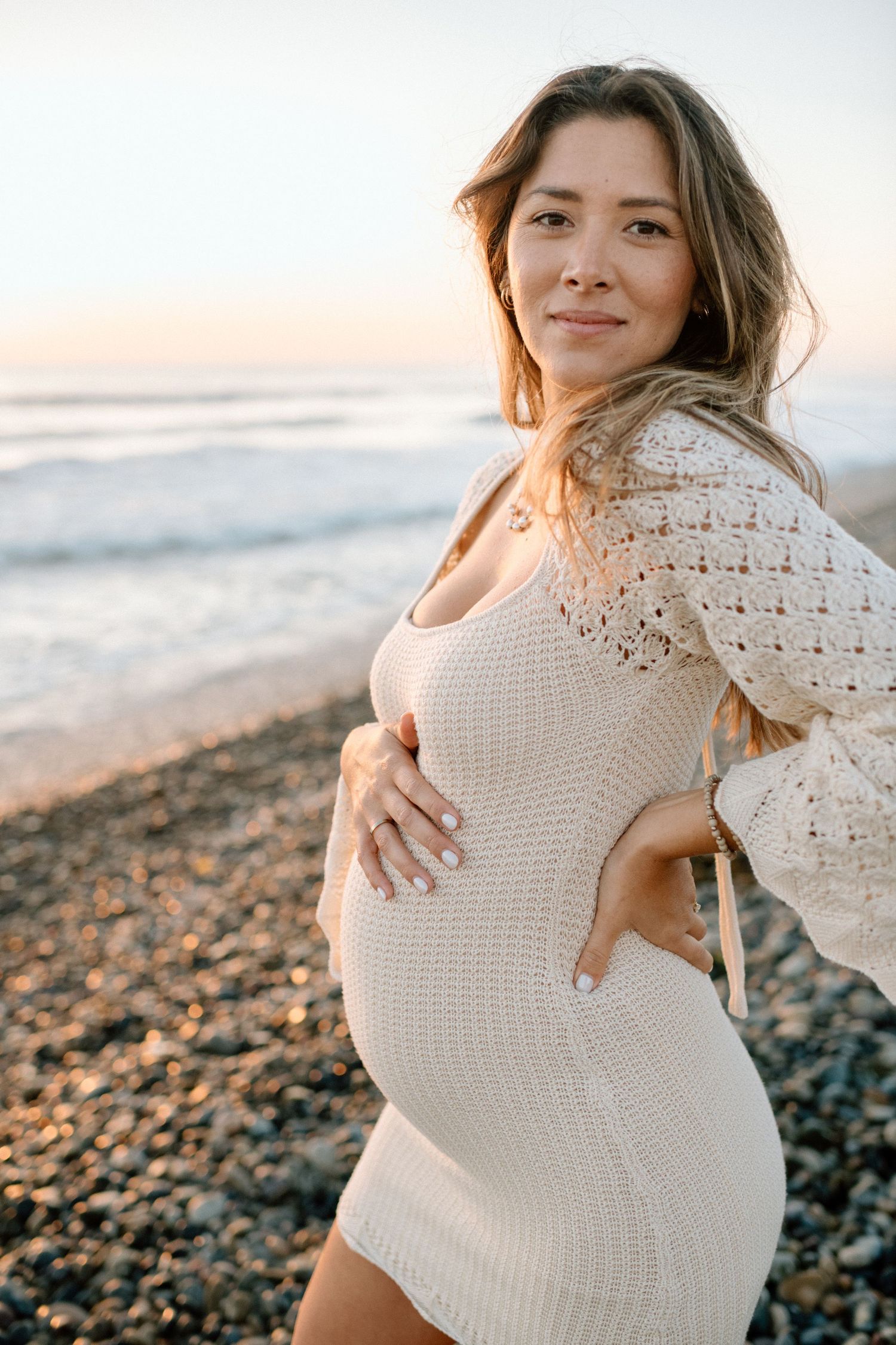 A person wearing a white knit dress poses on a beach at golden hour.