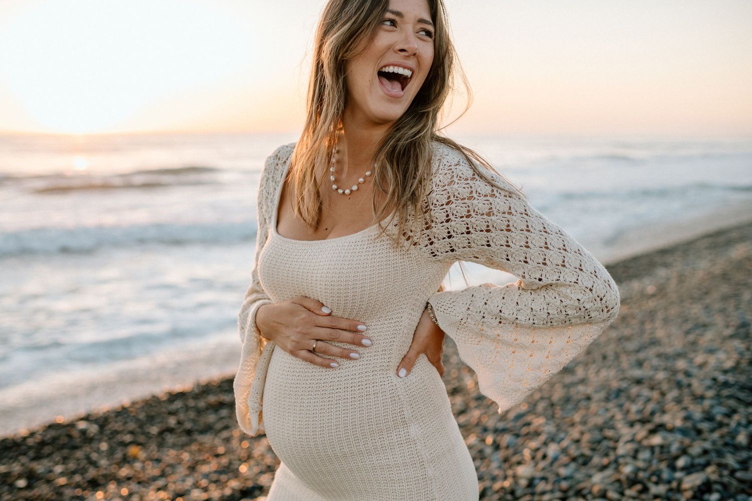 An expectant mother in a white dress poses on a beach during golden hour.